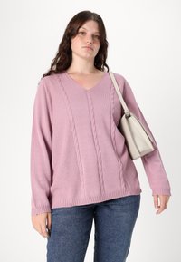 Pink knitted sweater with a V-neck, featuring textured cable patterns. Paired with dark jeans and a small light gray handbag.