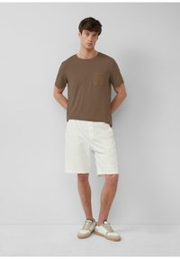 Braunes T-Shirt mit kurzen Ärmeln und einer Brusttasche, kombiniert mit weißen gerippten Shorts und beigen Turnschuhen mit hellen Akzenten. Einfaches Design.