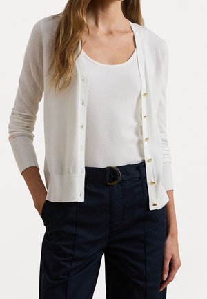 Vrouw draagt een wit gebreid vest met knopen over een wit hemdje, gecombineerd met hooggetailleerde donkerblauwe broek met riem.