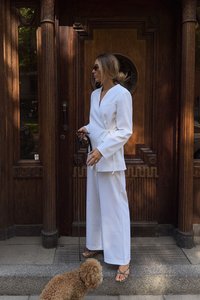 Traje blanco estilo envolvente con escote en V, piernas anchas y lazo lateral. La modelo lleva gafas de sol negras y sandalias con cintas, sujetando una correa. Fondo de madera marrón.