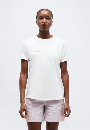 Femme debout, face à l'avant, portant un t-shirt blanc Mammut et un short violet clair sur un fond gris uni.