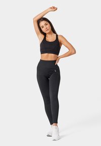 Melna sporta krūšturis ar apaļu izgriezumu un regulējamām siksnām, apvienots ar augstiem melniem legingiem ar tekstūru. Baltas sporta kurpes.