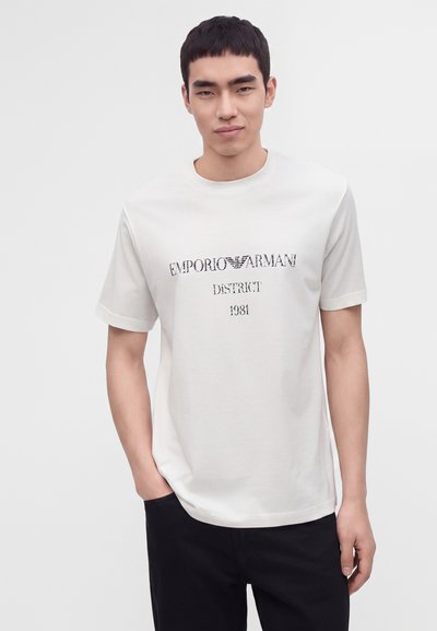 Νέος με κοντά μαύρα μαλλιά που φοράει λευκό t-shirt Emporio Armani και μαύρο παντελόνι, στέκεται με το ένα χέρι στην τσέπη.
