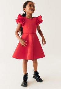 Robe rouge en tissu texturé, avec des manches courtes à volants et une jupe évasée. Le mannequin porte des bottines noires en cuir verni.