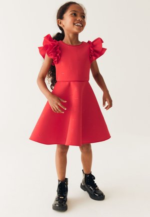 Robe rouge en tissu texturé, avec des manches courtes à volants et une jupe évasée. Le mannequin porte des bottines noires en cuir verni.