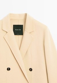 Manteau en mélange de laine beige avec un col cranté, une coupe à double boutonnage, des boutons noirs, et une étiquette noire portant la mention "Massimo Dutti".