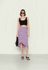 Saia envolvente roxa com padrão de leopardo preto, combinada com um top cropped preto. A modelo segura uma mala preta, usa óculos de sol escuros e sandálias de salto.