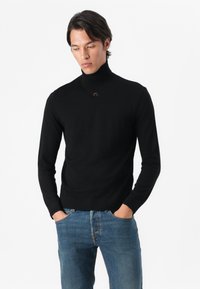Suéter negro de cuello alto hecho de una tela suave, con un corte ajustado y un discreto detalle de logo en el cuello, combinado con jeans de mezclilla azul.