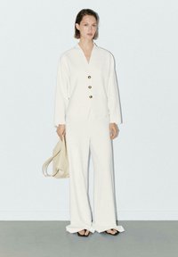 Veste blanche à coupe décontractée avec des boutons dorés et des manches longues, associée à un pantalon large. Les accessoires comprennent un sac beige et des sandales noires.