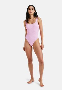 Vaaleanpunainen ribattua yhdestä palasta koostuva uimapuku, jossa on leveät hihnat ja neliömäinen pääntie. Uimapuvussa on istuva siluetti ja korkeat jalka-aukat.