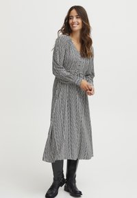 Juodai baltas raštuotas sijonas su V formos iškirpte, ilgomis rankovėmis ir plačiu apačiu, derinamas su iki kelių siekiančiais juodais batais.