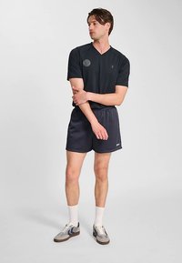 Svart kortärmad V-ringad tröja med en cirkulär logotyp, kombinerad med mörkgrå mesh träningsshorts och grå sneakers med blå detaljer.