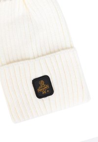 Bonnet en tricot couleur crème clair, avec une texture côtelée. Patch rectangulaire noir avec logo brodé sur un revers replié.