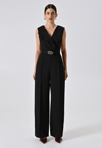 Tuta nera con design avvolgente, senza maniche e gambe larghe. Presenta una cintura coordinata con una fibbia dorata. Tessuto lucido e texturizzato.