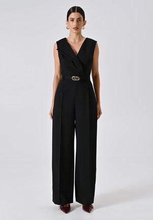 Combinaison noire avec un design croisé, sans manches et jambes larges. Équipée d'une ceinture assortie avec une boucle en or. Tissu brillant et texturé.