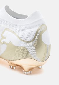 Λευκό και χρυσό ποδοσφαιρικό παπούτσι με πλεκτό επάνω μέρος, υφή στο πλάι, διαφανή τρακτέρ και εμφανές λευκό λογότυπο Puma στη φτέρνα.