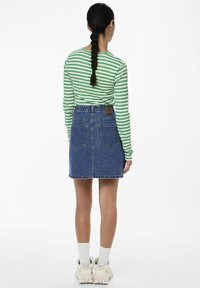 Nainen, jolla on lettiparta, pukeutuneena vihreä-valkoiseen raidalliseen pitkähihaiseen paitaan, sinisiin farkkuhameeseen, valkoisiin sukkiin ja valkoisiin tennareihin, kääntyy poispäin.