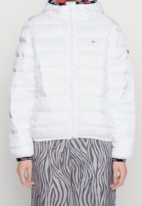 Vit puffjacka med dragkedja framtill och huva. Har horisontella quiltade paneler och sidofickor. Buren över grå zebra-mönstrade byxor.