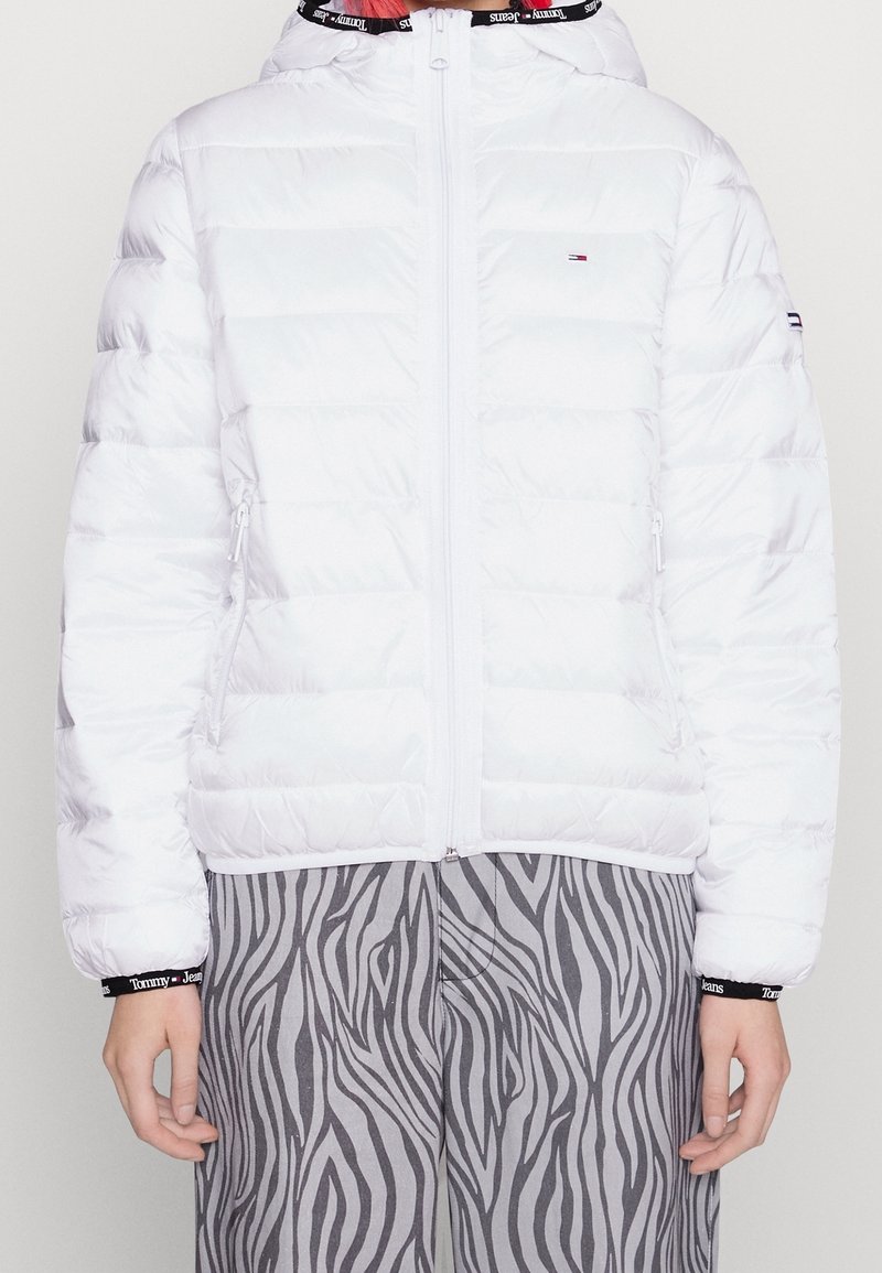 Vit puffjacka med dragkedja framtill och huva. Har horisontella quiltade paneler och sidofickor. Buren över grå zebra-mönstrade byxor.