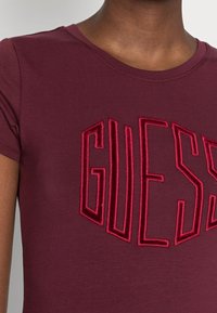 Nærbillede af en mørktonet person iført en maroon t-shirt med hævede, fløjlsbløde bogstaver, der staver "GUESS" på brystet.