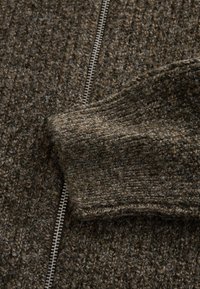 Bruin gebreid stof met een gestructureerd patroon, met een zilveren rits langs één zijde. Zachte en licht pluizige textuur.