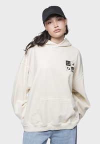 Sudadera de gran tamaño color crema, hecha de un tejido suave, que cuenta con un bolsillo frontal y un diseño gráfico con cuatro ilustraciones de ojos en el pecho.