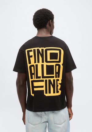 Άνδρας όρθιος με την πλάτη προς τα έξω, φοράει μαύρο μπλουζάκι με μεγάλα κίτρινα γράμματα που σχηματίζουν τη φράση "FINO ALLA FINE" και ανοιχτό μπλε τζιν.