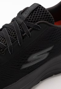 Scarpa sportiva nera con tomaia in rete, tessuto testurizzato e lacci a contrasto. Presenta il logo sul lato e una suola nera imbottita.