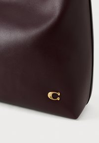 Borsa in pelle bordeaux con texture liscia, caratterizzata da un evidente logo dorato a forma di "C" sul lato. Forma curva e strutturata.