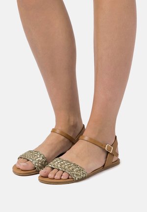 Piedi che indossano sandali flat color cuoio con cinturini intrecciati nella parte frontale e cinturini alla caviglia con fibbia su sfondo grigio chiaro.