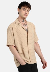 Camisa de manga corta de color beige claro con pequeños patrones de puntos multicolores, corte relajado y cuello con muesca, con botones en la parte delantera y textura suave.