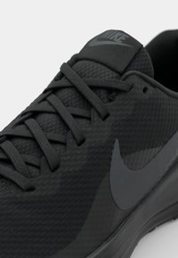 Svart atletisk sko tillverkad av texturerat syntetiskt material. Har platta snören, en vadderad plös och en framträdande grå swoosh-logga på sidan.