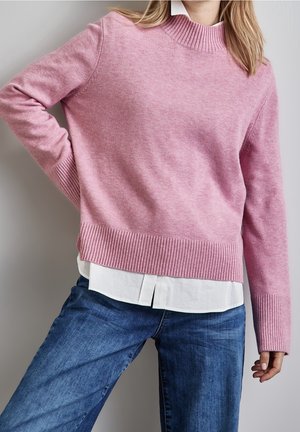 Personne portant un pull en maille côtelée rose par-dessus une chemise blanche et un jean bleu, debout, une main sur la hanche, devant un arrière-plan neutre.
