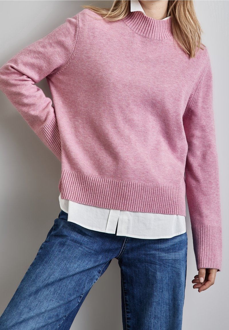 Personne portant un pull en maille côtelée rose par-dessus une chemise blanche et un jean bleu, debout, une main sur la hanche, devant un arrière-plan neutre.