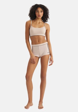 Mujer de pie descalza, vistiendo un top corto beige sin mangas y shorts de talle alto a juego con franjas laterales y un pequeño logo.