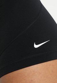 Shorts de sport noirs en tissu extensible avec des coutures flatlock et un logo Nike blanc sur le côté. Le matériau a une texture lisse.