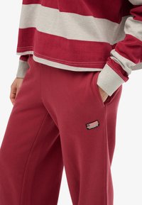 Person in rot-beiger gestreiftem Langarmshirt und roten Hosen, mit der Hand in der Tasche, die ein kleines Flaggenpatch auf den Hosen zeigt.