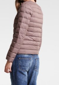 Polstret jakke i lys brun med horisontalt quiltet mønster, rund halsudskæring og tætsiddende silhuet. Sammen med blå denim jeans.