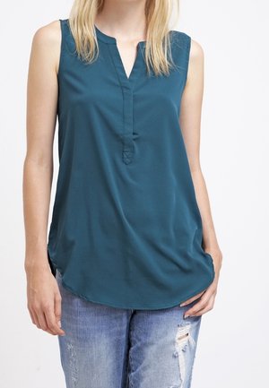 Ärmellose türkise Bluse mit geteiltem Ausschnitt, getragen zu hellblauen, abgenutzten Jeans an einer Person, die vor weißem Hintergrund steht.