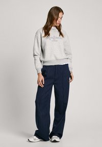 Sudadera gris con logo bordado, combinada con pantalones de pierna ancha a rayas azules. Modelo lleva zapatillas blancas y el cabello largo suelto.