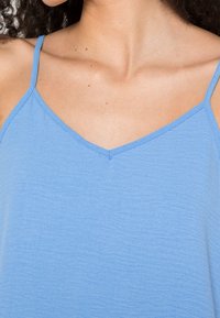 Débardeur bleu clair avec fines bretelles spaghetti, encolure en V, et tissu texturé. Vue en gros plan mettant en valeur la couleur et la forme de l'encolure.