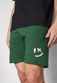 Pantaloni corti in cotone verde con una finitura testurizzata, featuring un sorriso bianco e un simbolo di croce. Fascia elastica in vita e design tapered.