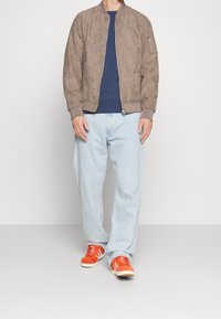 Veste aviateur en daim marron, t-shirt à col rond bleu marine, jean bleu clair et baskets orange avec accents blancs. Tenue décontractée en couches.