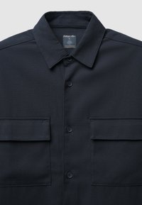 Camicia blu scuro con colletto strutturato, due tasche sul petto e un tessuto texturizzato. Presenta bottoni neri e un'etichetta all'interno.