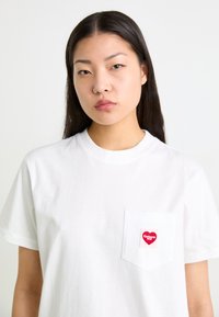 Bílý bavlněný tričko s krátkými rukávy, s náprsní kapsou zdobenou červenou a růžovou srdcovou nálepkou. Minimalistický design.