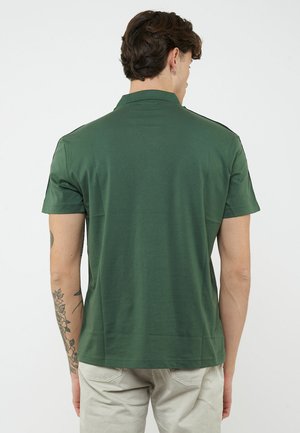 Man met tatoeages, gekleed in een donkergroen poloshirt met korte mouwen en lichtbeige broek, gezien van achteren tegen een effen witte achtergrond.