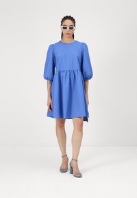 Kék, textúrált ruha, kerek nyakkivágással és puffos ujjakkal, deréknál beszorítva. Világoskék, tömbtalpú szandálokkal párosítva.