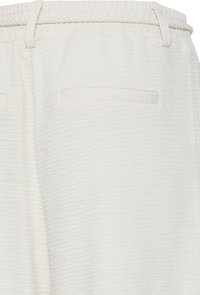 Gros plan d’un pantalon blanc texturé avec une taille élastique, des passants pour ceinture et une poche arrière horizontale sans fermeture visible.