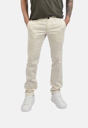 Uomo con braccia tatuate, pantaloni beige, camicia verde scuro, scarpe da ginnastica bianche, mani in tasca, in piedi davanti a uno sfondo semplice.