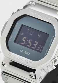 Montre Casio G-Shock en argent avec un affichage numérique rectangulaire, boîtier en acier inoxydable, verre teinté bleu et détail gravé "PROTECTION".